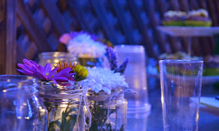 Tabletop with jars containing either tea light candles or flowers. A two-tiered cake stand with cupcakes is in the background on the right hand side.