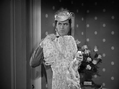 Cary Grant, wearing a hat, holding up a dress, looking in a mirror.