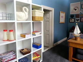 One-twelfth scale modern miniature lounge cube bookcase with various games and puzzles on the shelves along with a stack of magazines and a pair of binoculars.
