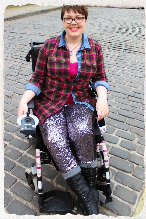 Young woman in an electric wheelchair, viewed head-on, wearing sparkly leggings, tall boots, and a plaid shirt.