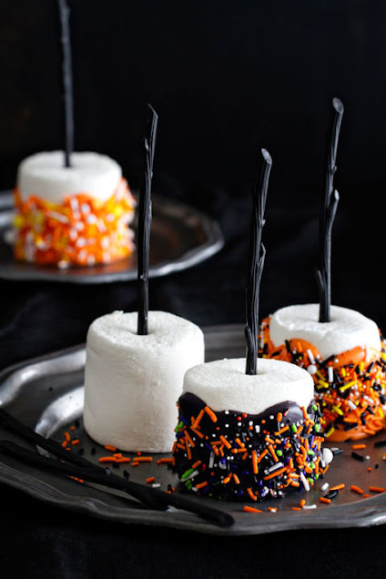 Marshmallows decorated with Halloween orange and brown sprinkles and a small pretzel stick in the marshmallows that looks like a witches broomsprinkles