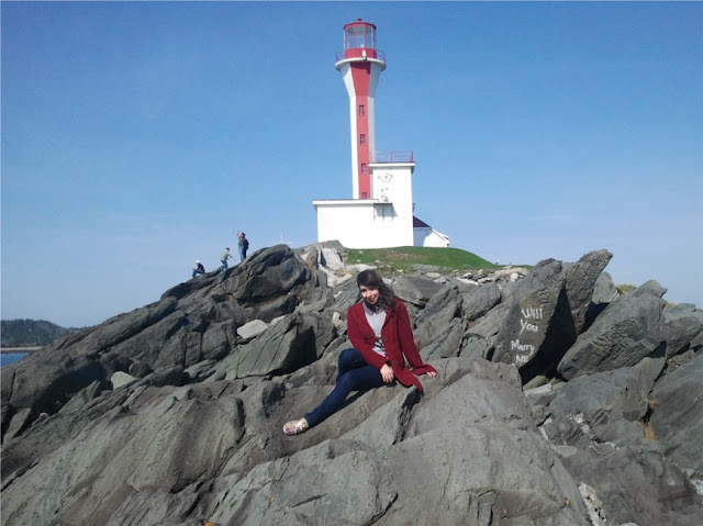Photo of Sam Routledge sitting on rocks with lighthouse in the background. Nearby rock has been spray painted with "Will you marry me?"