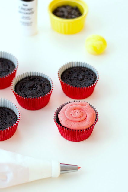 piping icing onto cupcakes chocolate cupcakes, one with red frosting swirl ready to be decorated