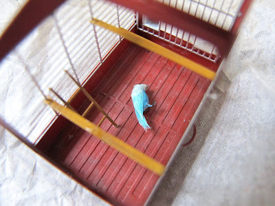 Modern dolls' house miniature bird cage, with a blue parrot on its side on the floor of it.