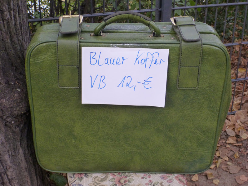 Grüner Koffer mit Schild "Blauer Koffer 12 Euro"