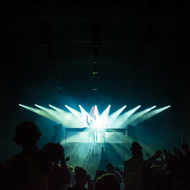 A dozen blazing spotlights focusing on a distant figure on a stage, framed by darkness and dark silhouettes.