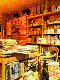 The right-hand side of a modern miniature shabby chic shop, showing wallpaper sample books, and shelves full of fabric and paint tins.