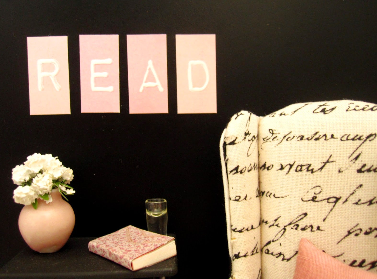 Modern doll's house miniature scene of a black and white wing chair against a black wall. On the wall are the letters R,E,A and D in pink and white and on the table under them is a pink vase with white flowers, a book and a glass of water.
