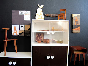 Modern dolls' house miniature display wall in a gallery. A collage art work is hanging on a black wall. In front of it is an art chair. On the left is a cabinet  with a wooden pear, fish and chair on top of it, and a table and wooden art work next to it. In the cabinet are a selection of items including a wooden ampersand tile, a modern tray, wooden bowls and a book safe.