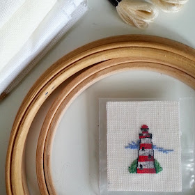 Miniature cross stitch picture of a light house, in the middle of a set of embroidery hoops, next to two skeins of embriodery thread and a pile of cross-stitch fabric.