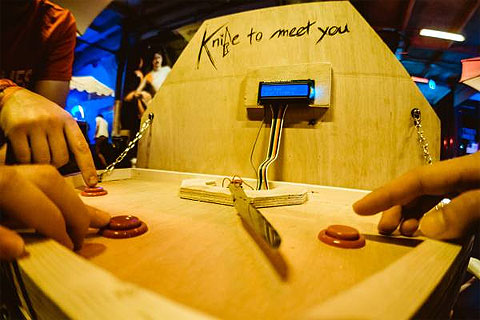 Photo of the Knife to Meet You game. A wooden game with an LCD display, rotating kitchen knife and three buttons spaced out in the knife's path with people nervously pushing them.