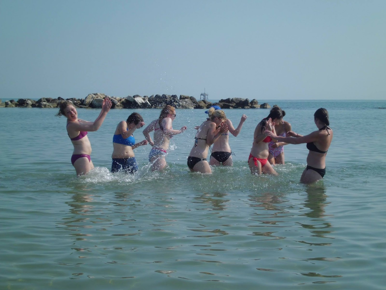 A group of women in bathing suits splash one another in the ocean.