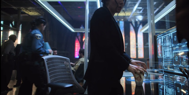 Ortega de pie, con la taza en la mano, ventanas tipo vidriera la fondo, pantalals luminosas con expedientes se reflejan en la mesa, gente de uniforme o sin el al fondo.