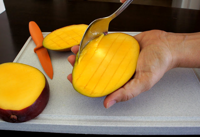 A hand holding a slice of mango using a spoon to scoop out the mango slices from the skin