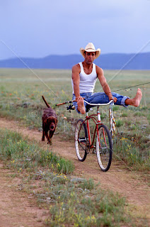cowboy-playing-around-on-a-bicycle-while-holding-fish-and-a-fishing-rod-on-a-ranch-with-a-dog-walks-along.jpg