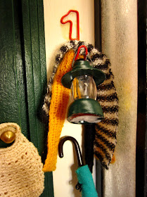 Modern dolls' house miniature wall hook holding two knitted scarves, a lantern and an umbrella.