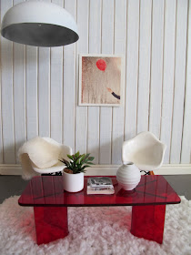 Modern one-twelfth scale miniature lounge scene with concrete floor, white panelled walls, white Eames chairs and a red perspex coffee table.