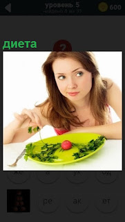 За столом девушка, перед ней тарелка с диетой, помидор и зелень