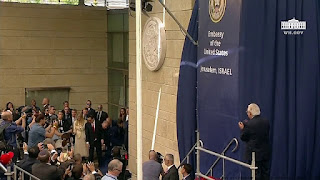 A photo of the press taking photographs and various dignitaries posing and applauding in front of a stone wall bearing a large stone seal of the US government. Next to the wall is a temporary banner reading 'Embassy of the United States, Jerusalem, Israel.'