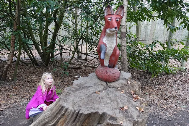 A girl in pink hiding behind a tree stump looking up at a wooden carved fox