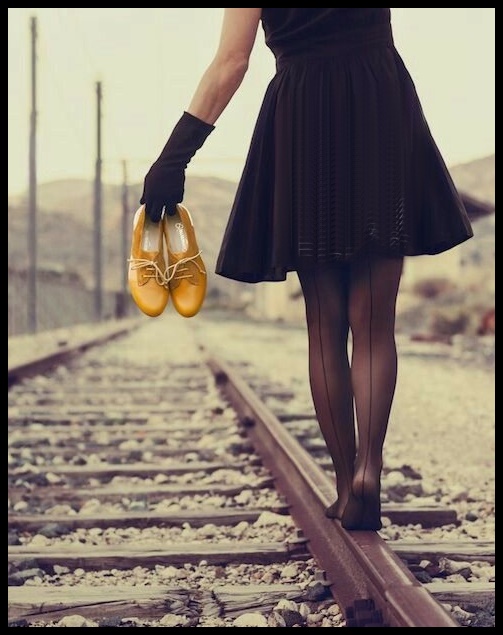 Uma jovem caminha descalça com os sapatos na mão, numa linha de trem.