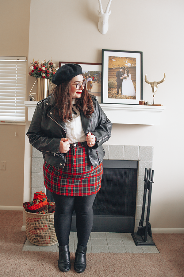 A plus size outfit consisting of a black beret, black faux leather moto jacket, white Peter Pan collar blouse, classic red tartan plaid mini skirt, black tight and black heeled chelsea boots