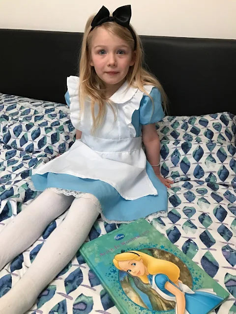 A 5 year old girl in a pale blue dress with white apron and white tights and a black hairband sitting next to a Disney Alice In Wonderland book