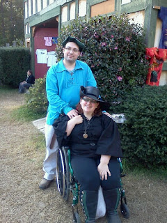 Author seated in wheelchair wearing black leather boots, black leggings, black pirate shirt, & black leather hat, with partner wearing khaki pants, turquoise pirate shirt, and green tam.