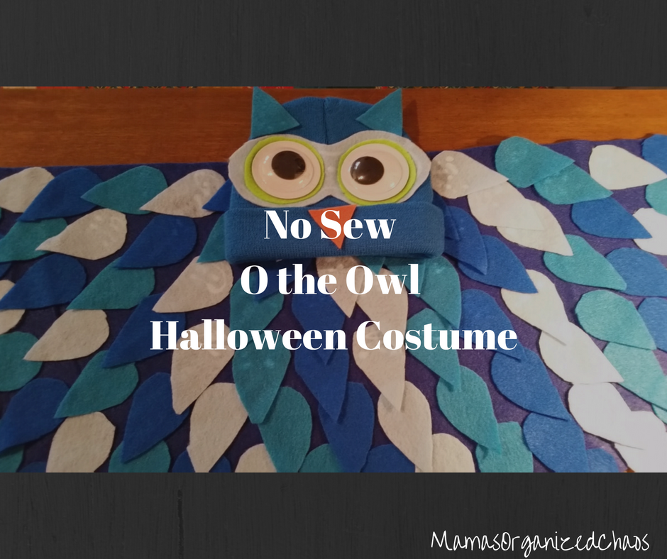 Blue ski hat turned into an owl with big google eyes and an orange nose, lying on a blue sweatshirt that has teardrops added to look like owl feathers for an O the Owl Halloween costume.
