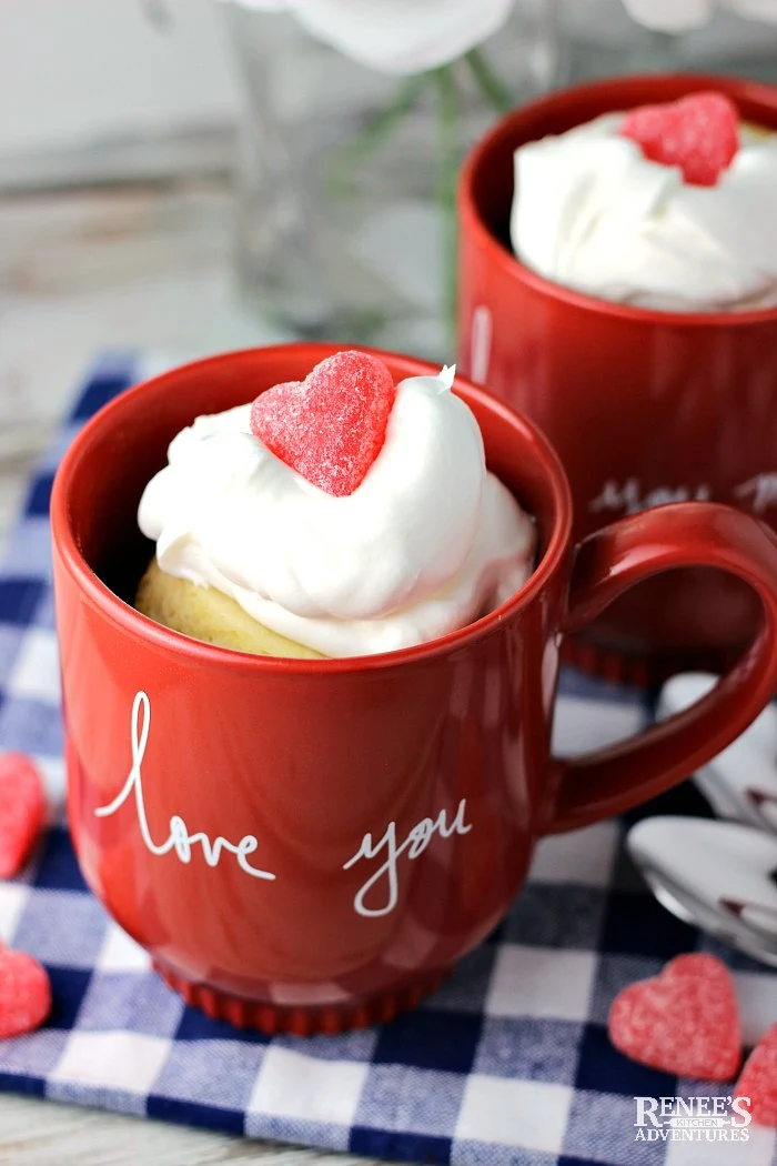 Two Vanilla Cake in a Mug on a blue and white checked cloth with whipped topping and gummy hearts