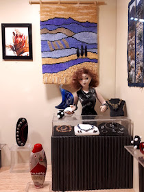 One-twelfth scale modern miniature art gallery, with a woman standing behind a counter in the corner with a display of jewellery in it. On the wall behind her is a tapestry and a painting and in front and to the side of the counter are a selection of colourful glass vases.