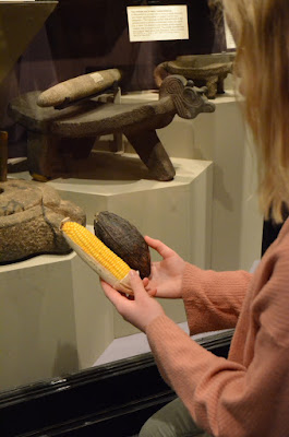 Girl holds a cocoa pod and a corn on the cob in her hand, sitting infront of a metate (a grinder)