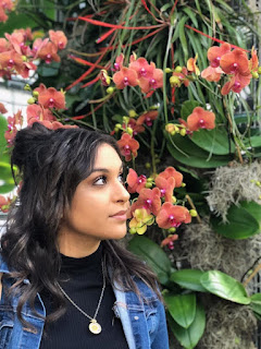 Photo of Neveen Shawish. Image of Neveen Shawish standing in front of a large orchid with salmon colored blossoms. She looks at the flowers, and is wearing a black, mock turtle neck top with a jean jacket, and a large gold pendant necklace. She has long, wavy chestnut brown hair and brown eyes.