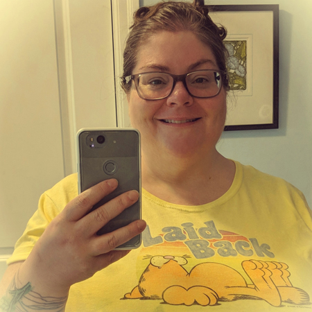image of me from chest up in a mirror, with my hair up and wearing grey-framed glasses, wearing a yellow t-shirt that has Garfield the Cat on it and text reading: 'Laid back.'