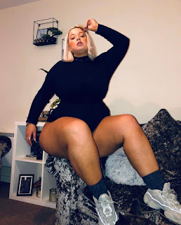 Plus size woman with blonde hair sitting on arm of sofa wearing black bodysuit and trainers