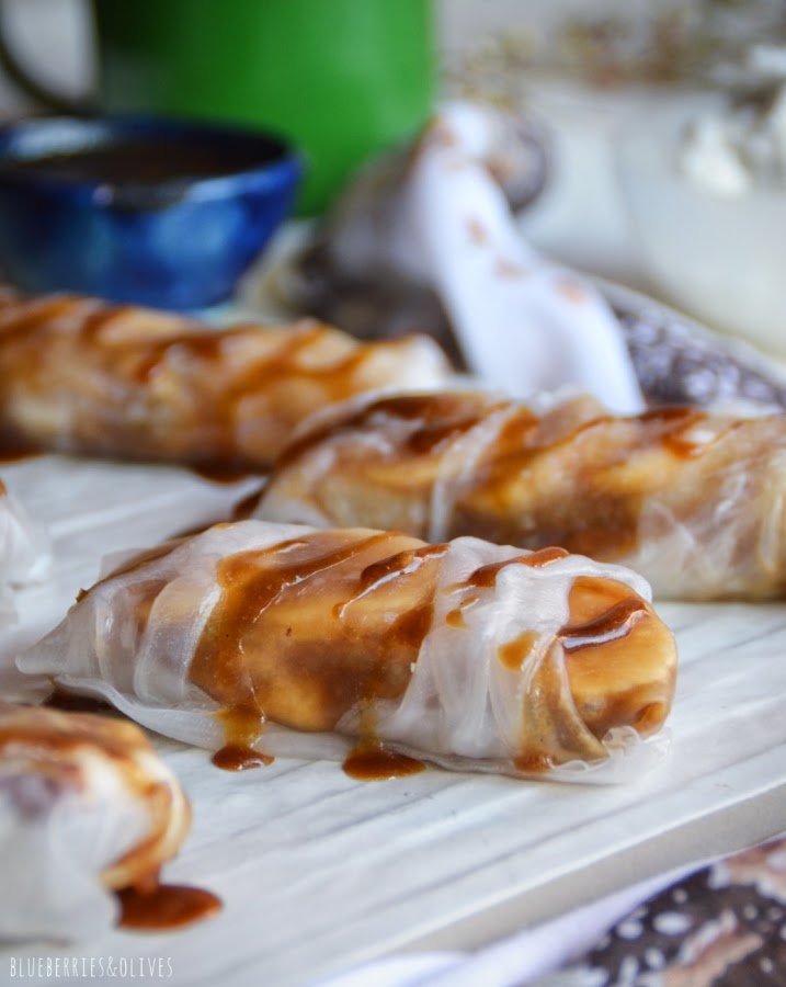 ROLLITOS DE PLÁTANO Y CARAMELO EN SUPERFICIE BLANCA, FONDO CON FLORES SECAS Y TAZA VERDE