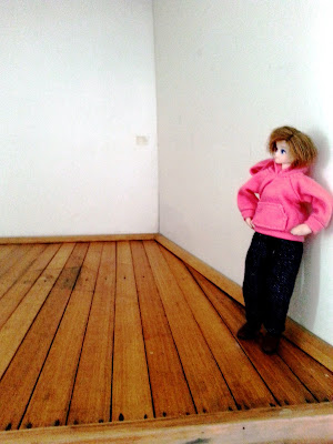 Modern dolls' house miniature doll standing on a wooden floor and leaning against a white wall.
