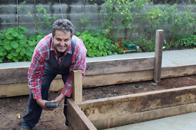 Brendan Murphy stands beside a raised wooden garden bed. The bed is only partly constructed and he is holding a drill to wood in the corner as he looks and smiles at the camera.
