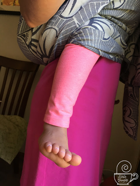 [Image from the side of a child worn on her Mama's back in a grayish purplish geofloral woven wrap carrier. Mom is wearing a pink dress. Child has on peachy orange leggings. Her legs are so long that Mama has a heard time believing it.]