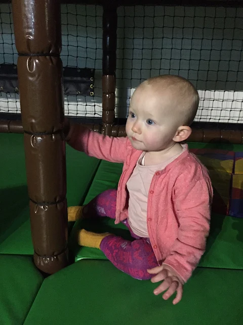 A nearly one year old girl wearing shades of pink (and mustard yellow socks) sitting on a green mat with a padded brown pole in front of her
