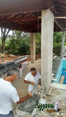 Bizzarri ajudando a fazer o revestimento com pedras do rio, sendo esse revestimento de pedra nos pilares de concreto. Pedra do rio no tamanho de uma laranja.