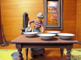 Model of a 19th-century woman inside a house, pouring milk from a jug into several bowls.