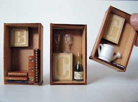 Three miniature cedar crates containing letters of the alphabet and items that match. From left to right: 'B' and five miniature books, 'C' and a bottle of champagne with two glasses, and 'G' with a watering can and trowel. A finger is shown for scale.