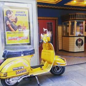 Modern dolls' house Vespa scooter parked in front of a cinema lobby.