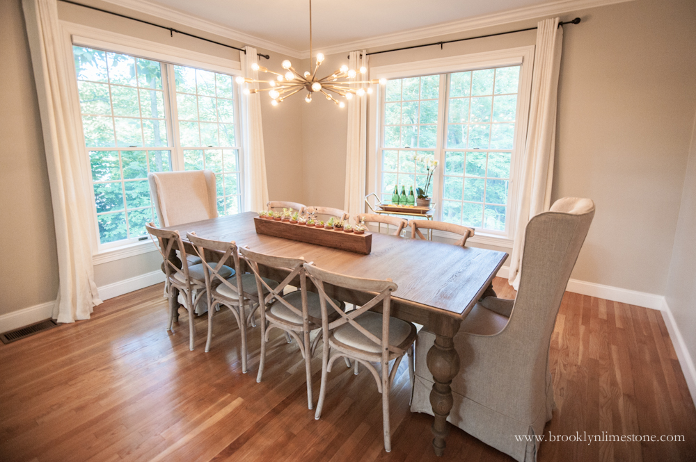 Dining room with cream curtains on 2 double hung windows, new grey wood table with upholstered cream end chairs and 3 cross backed chairs on each side