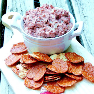 Braunschweiger Dip in a white bowl on a cutting board with pretzel chips on a wooden background with a red and white checkered napkin.