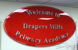 Welcome to Drapers Mill Primary Academy chrome vinyl letters on a red oval perspex sign mounted to the reception wall.