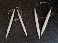 Two sets of grey plastic circular knitting needles on a dark background.  The cables are coiled at the top and the needle ends are pointing down. The set on the left are smaller and thinner than those on the right: 12 mm and 15 mm wide respectively. 