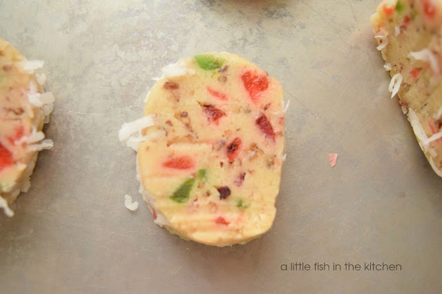 Freshly prepared Santa's Whisker cookie dough is sliced and read to bake on a parchment paper-lined baking sheet. The uncooked cookie slices are placed on a baking sheet two inches apart. Colorful bits of candied cherries and pecans are visible in the cookie dough and the fringe of white sweetened coconut shreds is around the edges. 