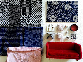 A flat lay with three pieces of blue and white Japanese-patterned fabric (and one in red), a card with a piece of blue and white japanese fabric on its front, a deep red miniature velvet sofa, a one-twelfth scale Yanagi butteryfly stool, three one-twelfth scale lucky cats, two one-twelfth scale cherry-blossom cushions, a pair of one-twelfth scale slippers and two pairs of one-twelfth scale chop sticks. All on piece of white woven wallpaper.